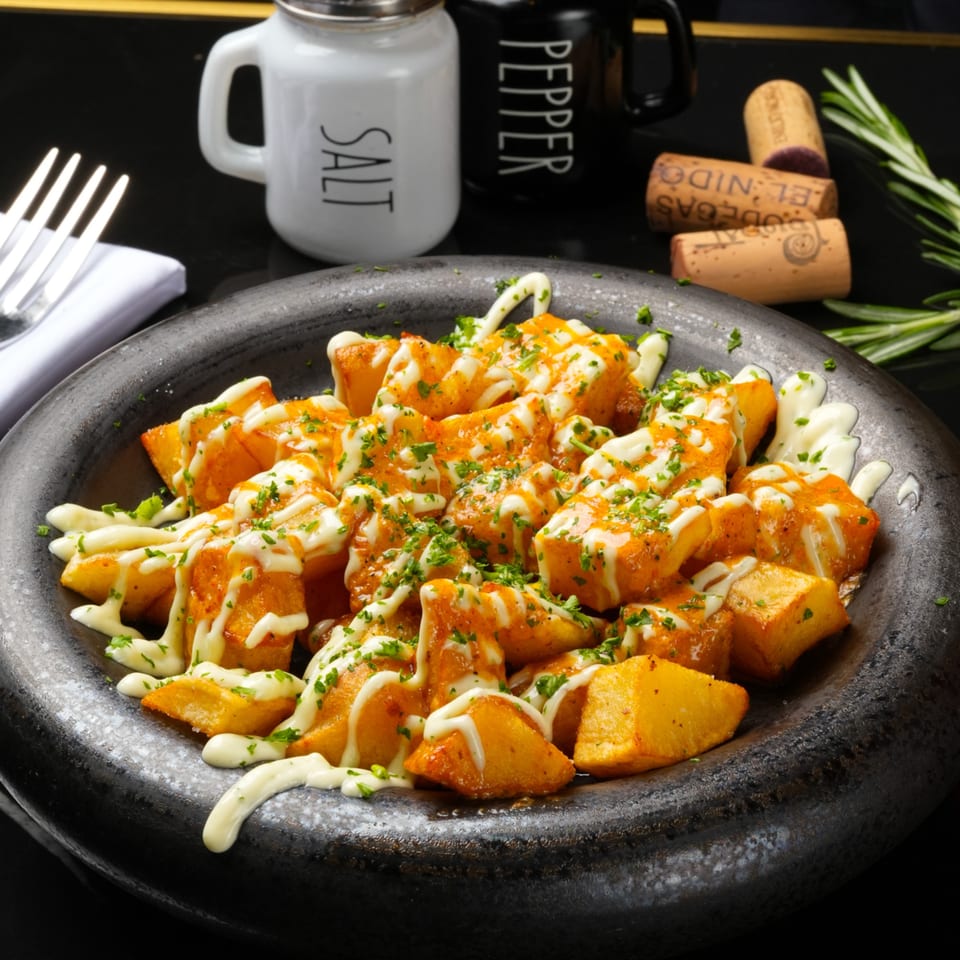 Patatas bravas, fried potato cubes with spicy sauce and aioli, garnished with parsley in a dark bowl.