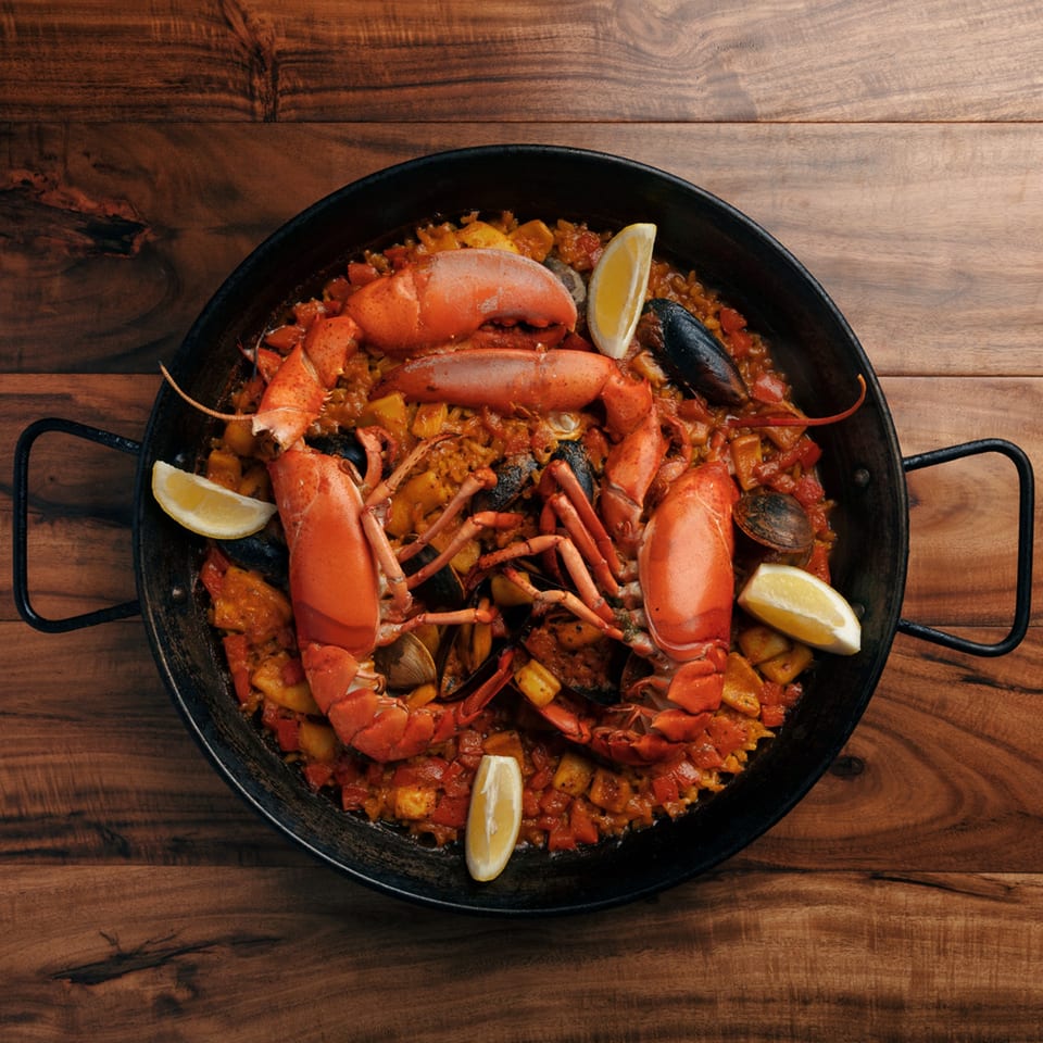 Xixon's Spanish paella with whole lobsters, mussels, clams, and lemon wedges in a black pan.
